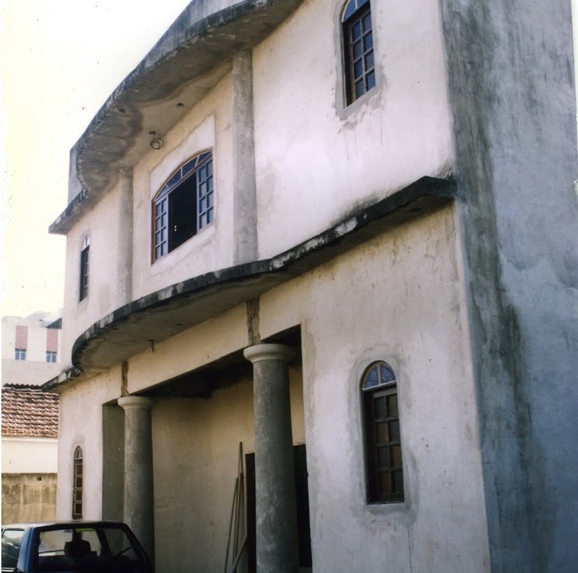 Fachada da obra em dois pavimentos, ainda sem pintura. Duas colunas cilíndricas sustentam a varanda curva do pavimento superior, e as janelas têm arco pleno com esquadrias azuis.