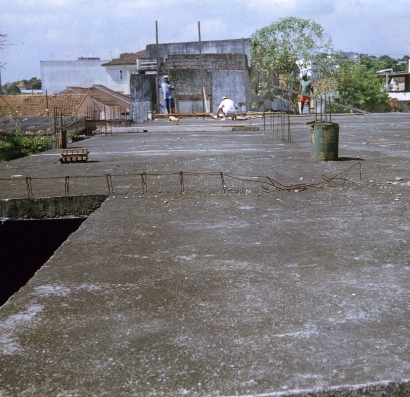 Grande laje de concreto já curada, vista rasante. Ao fundo, três trabalhadores e, além deles, os telhados e as casas do bairro.