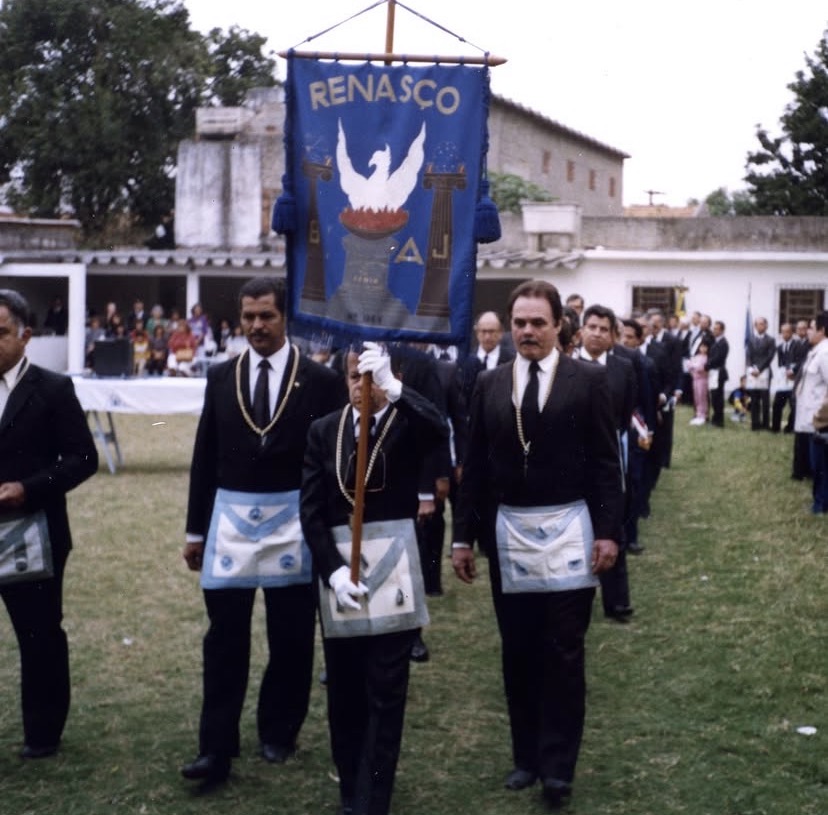 Cortejo maçônico ao ar livre. À frente, um irmão de luvas brancas porta o estandarte azul da Loja, com a palavra RENASÇO em ouro e a Fênix branca sobre as chamas. Ao lado dele, irmãos de terno escuro, colar e avental caminham pelo gramado.