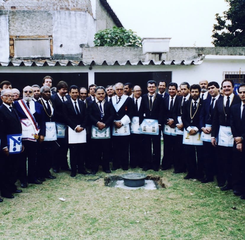 Cerca de trinta irmãos perfilados em semicírculo no gramado, de terno escuro, colar e avental maçônico. Em primeiro plano, a cova aberta com a pedra fundamental já assentada.